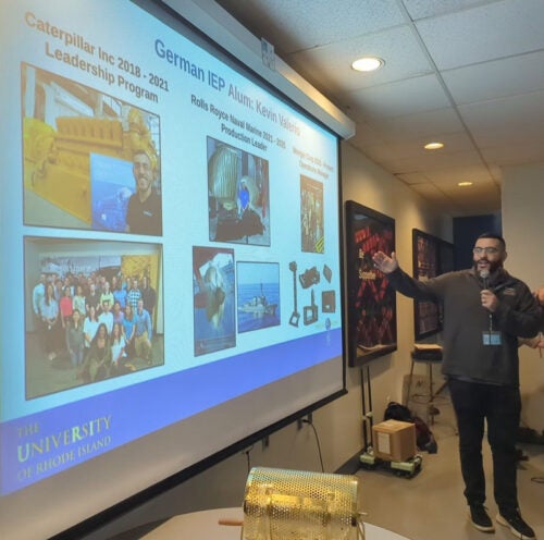 Kevin Valerio stands in front of a slide showing his career experiences since he completed the IEP, starting with Catepillar, to Rolls Royce and now Wenger Corp.