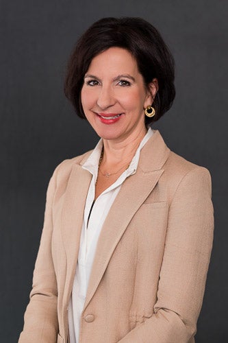 A formal portait of Kristen Riley, wearing a tan bllazer and white shirt on a gray background
