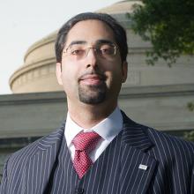 A headshot of Nevan Hanumara wearing a striped suit and red tie posed in front of a building.