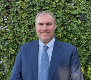 A headshot of Ryan Light wearing a blue suit, shirt and tie in front of a background of ivy.