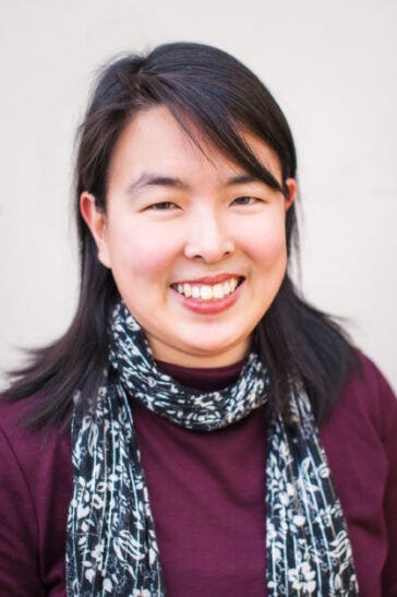 A headshot of Sarah Koenig wearing a maroon shirt and floral print scarf in front of a light neutral background.
