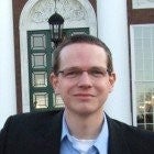 A headshot of Tobias Lührig wearing a dark suit in front of the entryway of a building with columns.