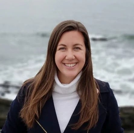 Professional headshot of a smiling woman with long brown hair wearing a white turtleneck and dark navy blazer, photographed against a blurred ocean and waves in the background.