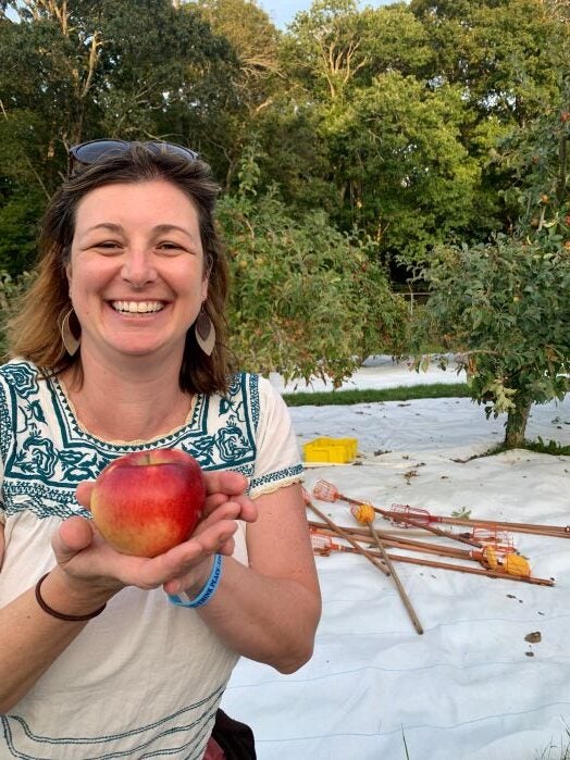 A smiling woman with dark hair wearing a decorative blue and white patterned shirt holds a fresh red apple in her hands while standing outdoors. Behind her is a scenic waterside setting with lush green trees, calm water, and driftwood along the shoreline.