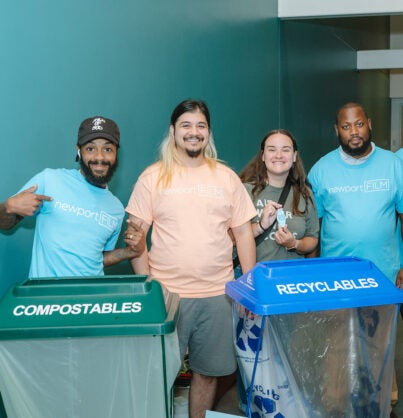 Four smiling volunteers wearing Newport FILM branded t-shirts pose together in front of a turquoise wall with a green composting bin and blue recyclables bin. The group represents an environmental or community recycling initiative, with three people in light blue shirts and one in a pink shirt.
