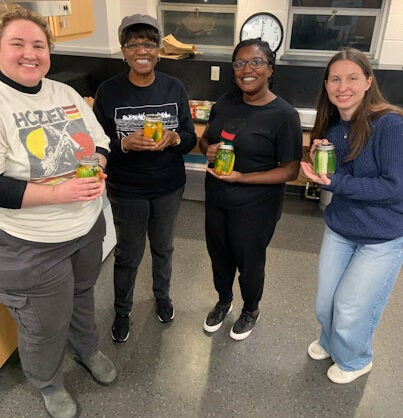 Four smiling young people stand together in a kitchen holding packaged and canned food items. They appear to be participating in a food-related program or event, with kitchen cabinets and windows visible in the background.