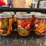Six glass mason jars with metal screw-top lids containing homemade pickled vegetables arranged on a dark speckled countertop in a kitchen. The jars display colorful preserved vegetables including red peppers, green peppers, and mustard seeds in golden-amber brine, with kitchen cabinets and appliances visible blurred in the background.