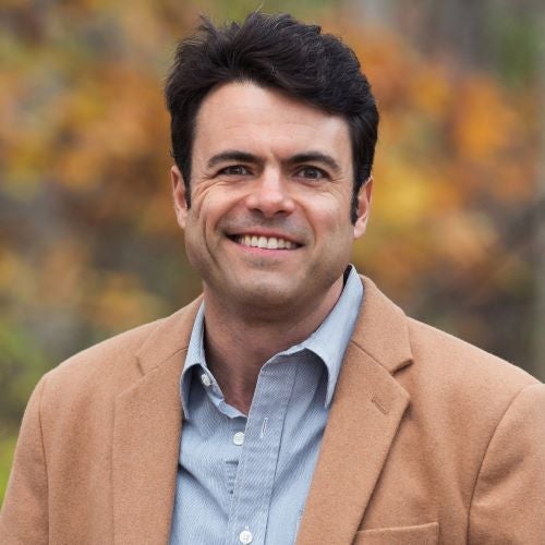 Christopher Riely, wearing a light brown sports jacket and light blue collared shirt, smiling, with a yellow bush in the background.