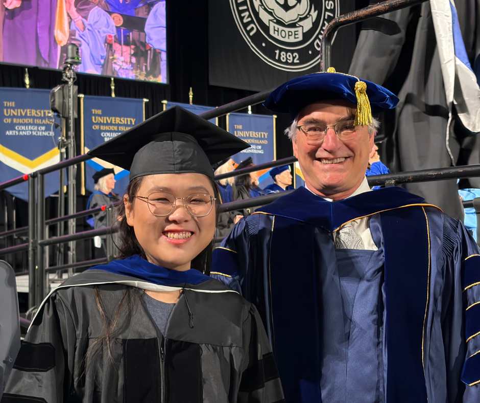 A student and faculty member smile in the Ryan Center, in their caps and gowns. 