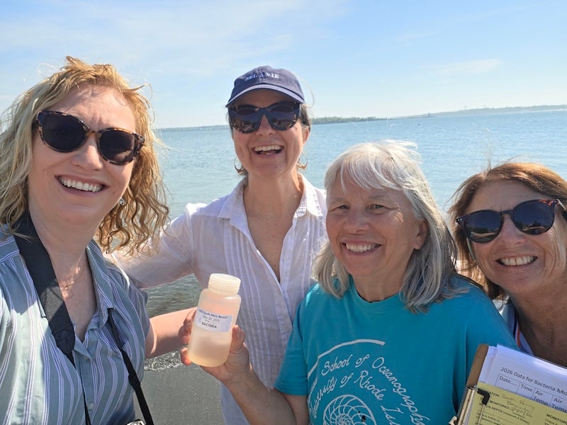 Four women smiling at the camera while outside on a beach on a sunny day. One is holding a water sample bottle.