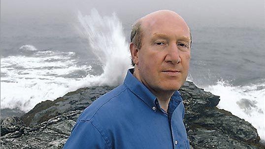 Headshot of Isaac Ginis, who is tanding outside with a wave hitting the rocky shore behind him.