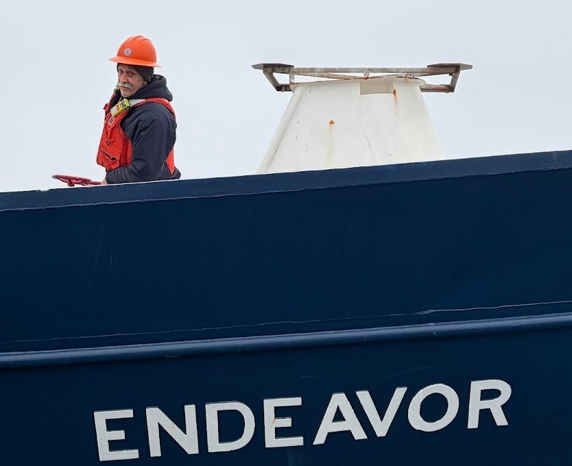 Kevin Walsh aboard the research vessel Endeavor. he is wearing a hard hat and safety vest, and the name of the vessel is printed on large letters on the hull below him