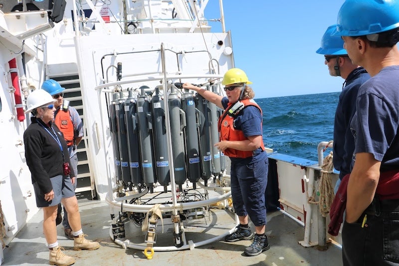 Lynne Butler aboard R/V Endeavor, wearing a hard hat and safety vest outside on the deck of the ship. She is showing a group of four educators in safety helmets how an oceanographic instrument works.