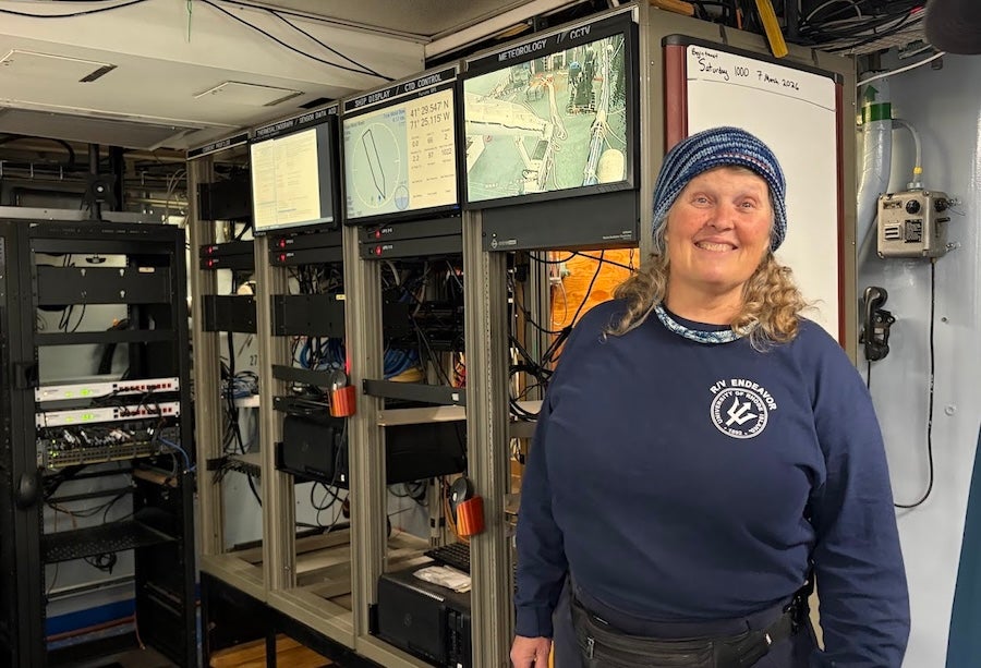 Lynne Butler smiling and looking towards the camera. She is standing inside the Endeavor in front of a series of computer monitors and technical equipment.