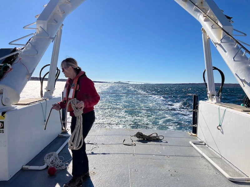 Lee Singer on the stern deck of the Endeavor. She is wearing a safety vest and managing ship lines under a large crane. The ship's wake is visible behind her, as is a bridge in the distance.