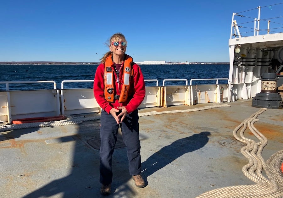 Lee Singer smiling at the camera on the back deck of the research vessel Endeavor. She is wearing a safety vest and is in calm seas with land far in the background.