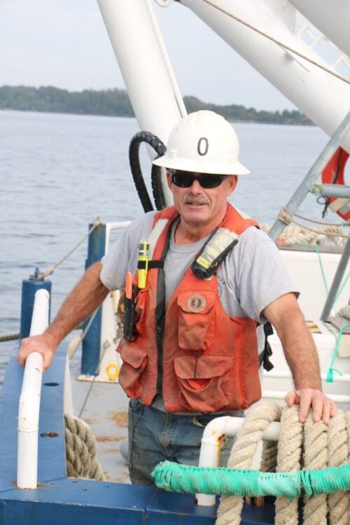 Oscar Sisson standing on the stern of a ship, wearing safety vest, hard hat and sunglasses.