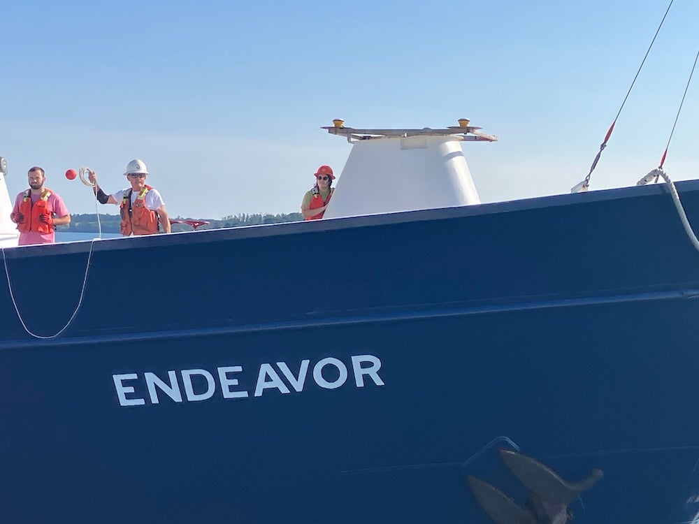 View of the research vessel Endeavor during a sunny day. Three people are on board, including Oscar Sisson who is preparing to cast a line to the pier.