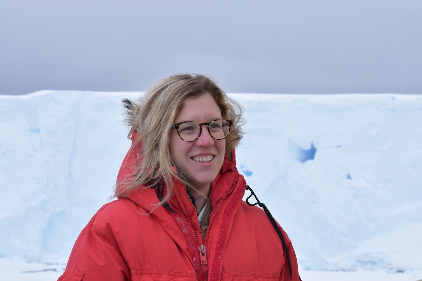 Headshot of Olivia Ahern, she is wearing glasses and a red winter jacket outside with Antarctice ice and an overcast sky in the background.