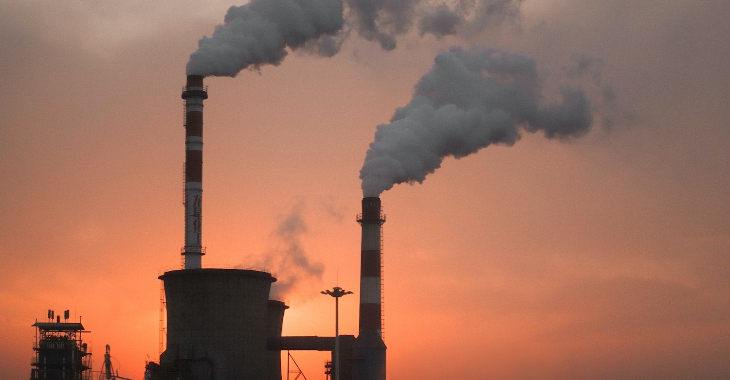 Two factory smokestacks with smoke coming out of the top set against a orange sky at sunset.