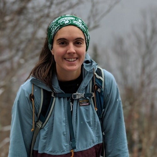 Headshot of Ali Johnson, seen outside with trees behind her, she is wearing a rain jacket and backpack.