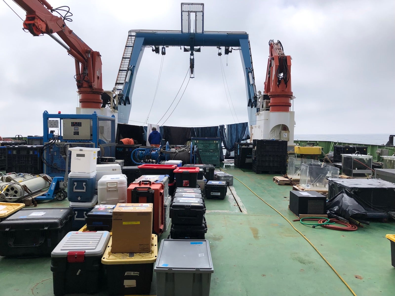 A large number of crates and boxes on the back deck of a research vessel, the deck is green and three large cranes are behind the collection of boxes and crates.