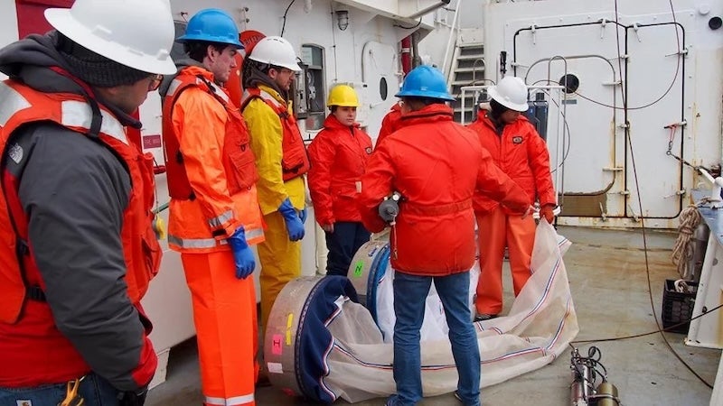 Seven students, each wearing hard hats and bright safety clothing, aboard a research vessel standing next to two large nets used for ocean research.
