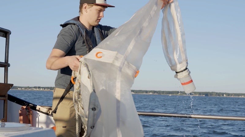 A student holds a large, fine-screen net aboard a boat in coastal waters.