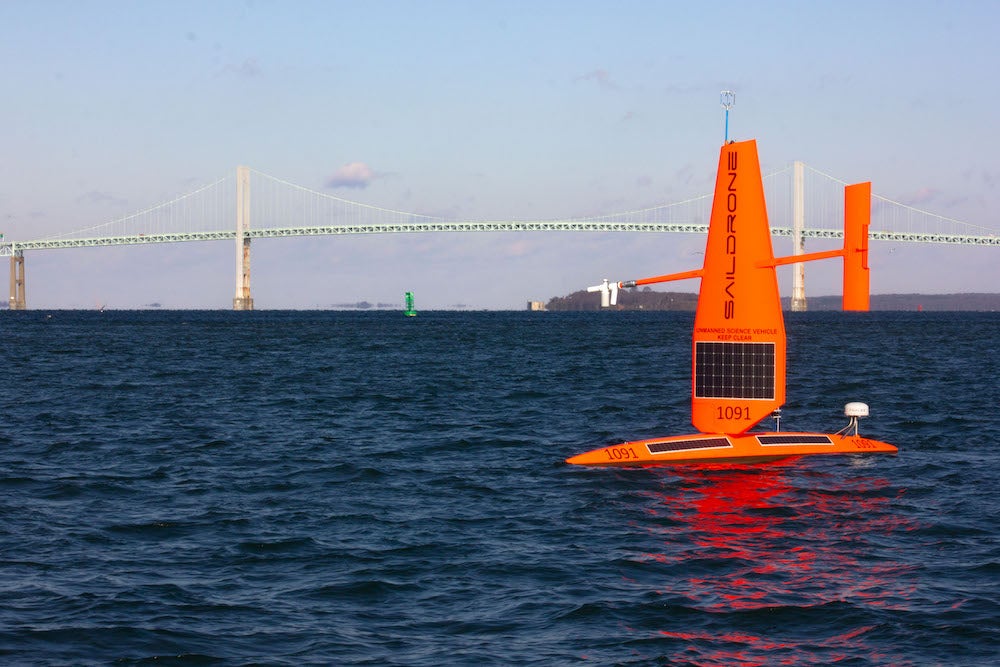 A bright orange saildrone, which looks like a robot sailboat, sails on the ocean with the New port Bridge in the background