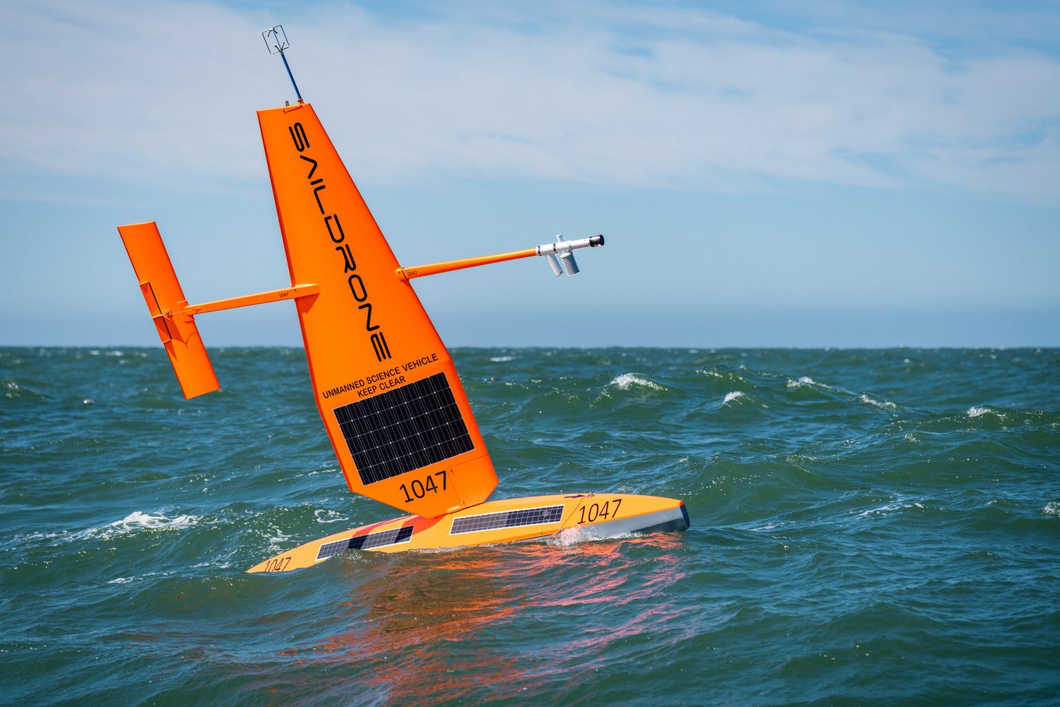 A Saildrone sails in rough seas. The drone is a bright orange boat without any people and has numerous scientific instruments attached to its sail made of hard materials (instead of fabric). .