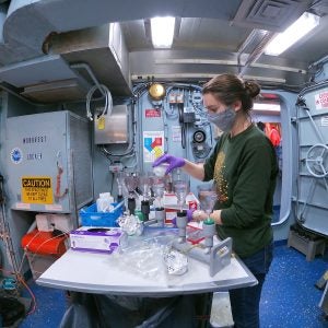 Female student working in a lab aboard R/V Endeavor, seen from the side and puring liquids into containers. She is wearing a face mask.