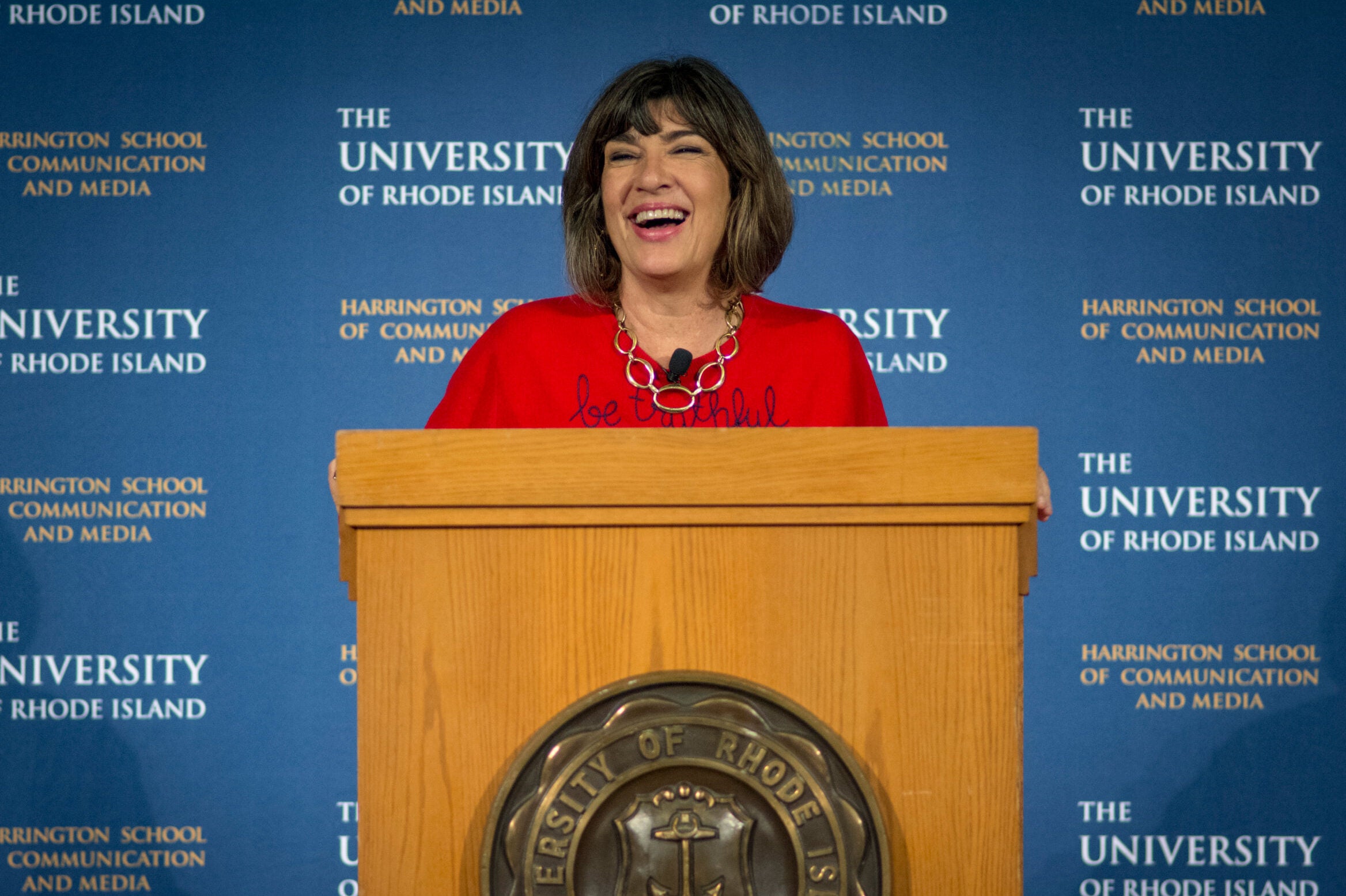 Christiane Amanpour standing at a lecture, laughing, with the University of Rhode Island logo on the backdrop.