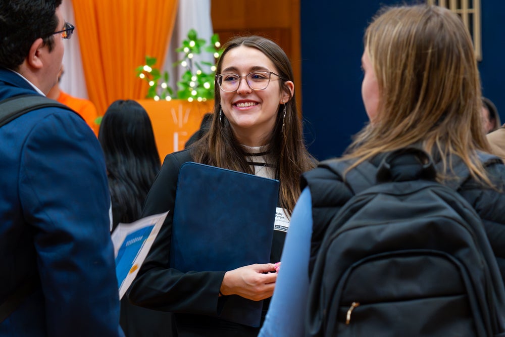 A student smiles while speaking with attendees at the Harrington School career fair, holding a portfolio folder as she connects with professionals and peers during the event.