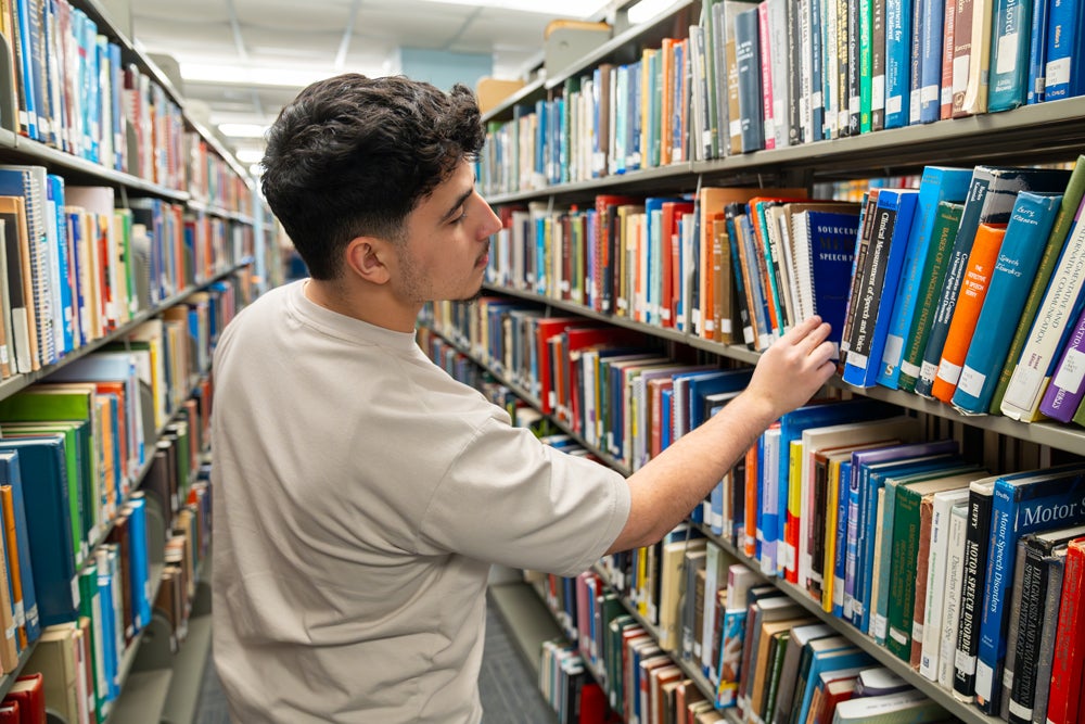 A student browses books on a library shelf, reaching for a book while standing between rows of shelves filled with academic texts.