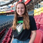 2022 alum, Siobhan Richards, posing in a stadium in a tshirt with her company's logo on it.