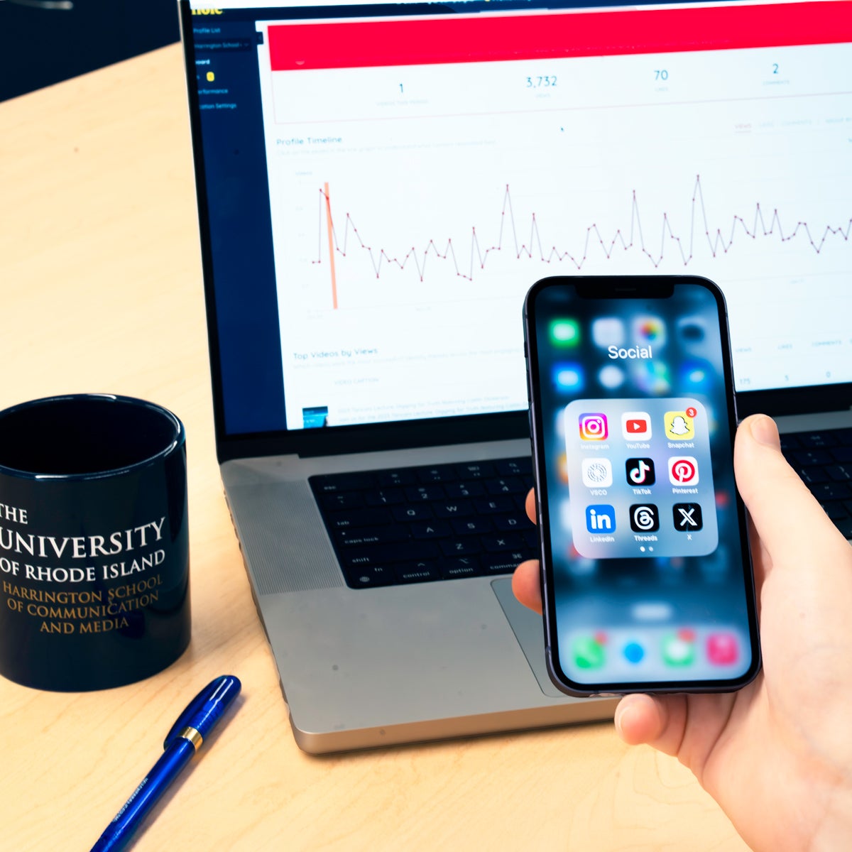 student holding an iphone with social media apps open in front of a computer with analytics on the screen