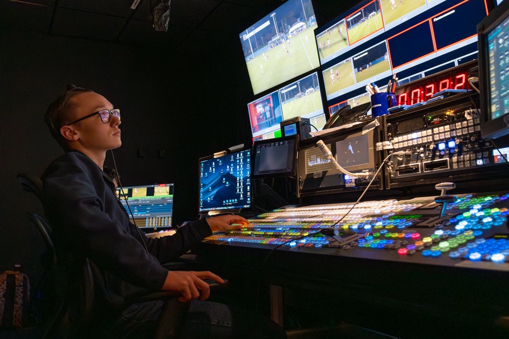 A student operates a broadcast control panel in a production booth, monitoring multiple screens showing a soccer game feed while managing live video equipment.