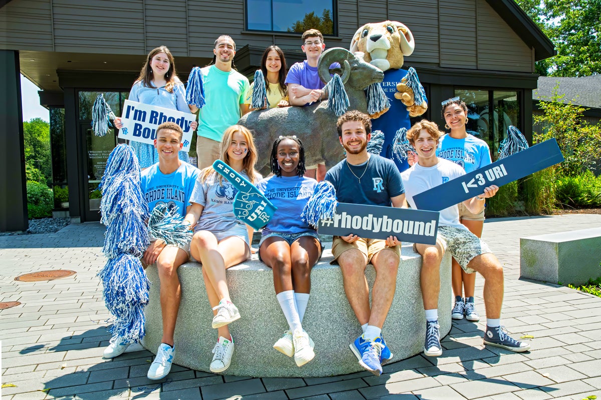 Students sit with the ram in front of the welcome center with signs about getting accepted to URI