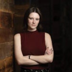 Caroline McCullough, a journalism and anthropology student, poses for a portrait wearing a burgundy sleeveless top against a dark background with a wood-paneled wall.
