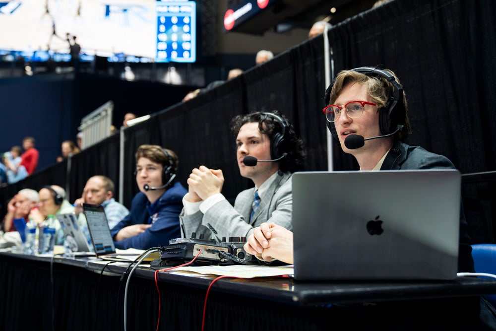 Students wearing headsets work at a broadcast production table with laptops and audio equipment during a sports media event, coordinating coverage from the sidelines.