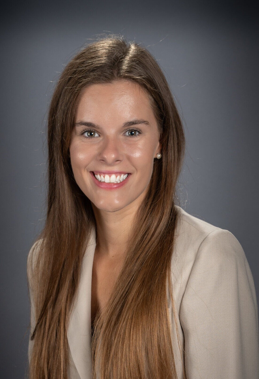 Headshot of Abigail, a young woman with chest-length brown hair, wearing a light brown blazer. She is smiling softly and facing the camera.