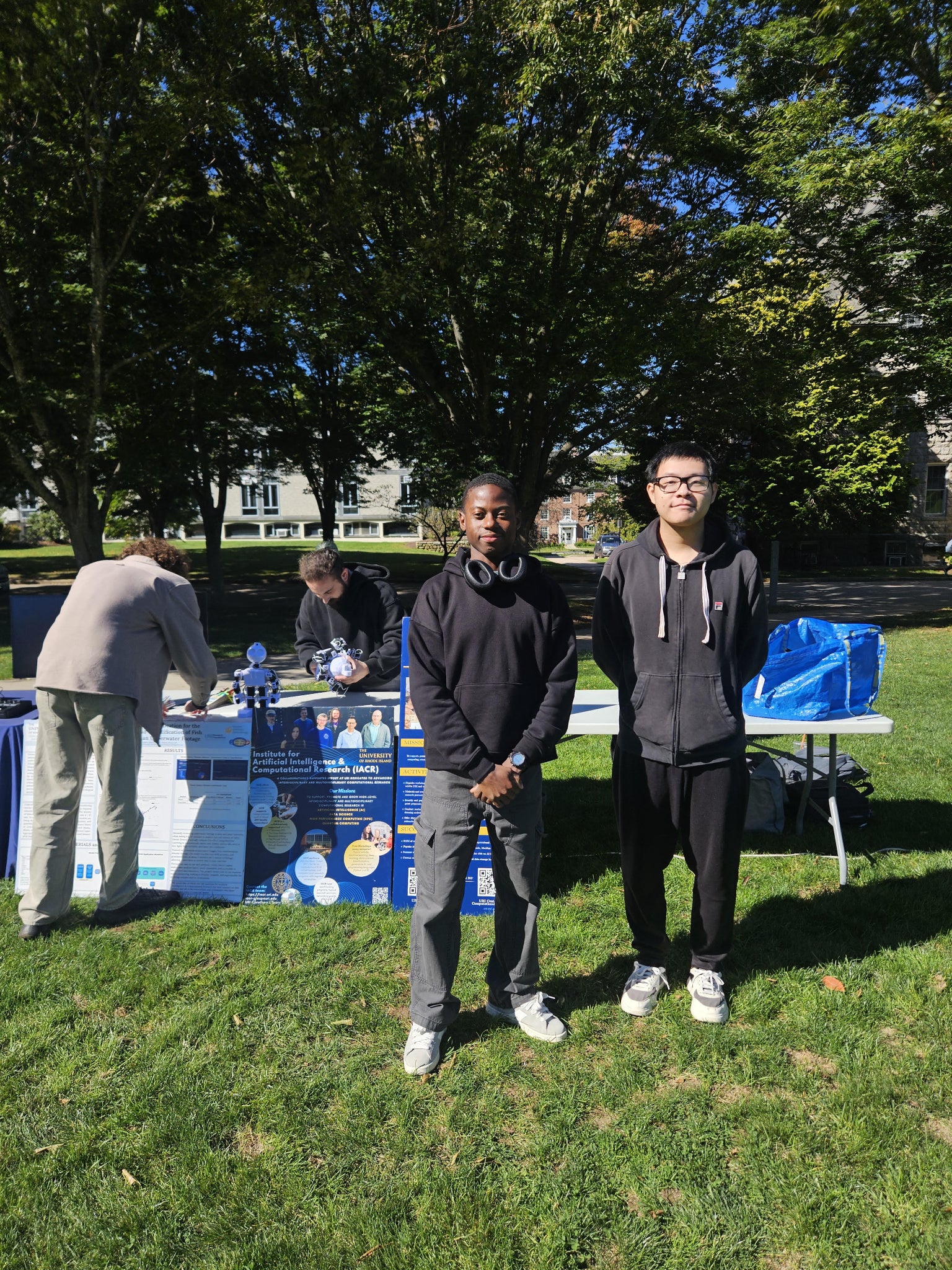 Students beside an IACR outreach table with research posters and a small robot on the URI campus.