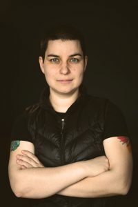 Photo of White woman with short, black hair, wearing a black vest and black t-shirt. She appears to be standing with her her arms are folded.