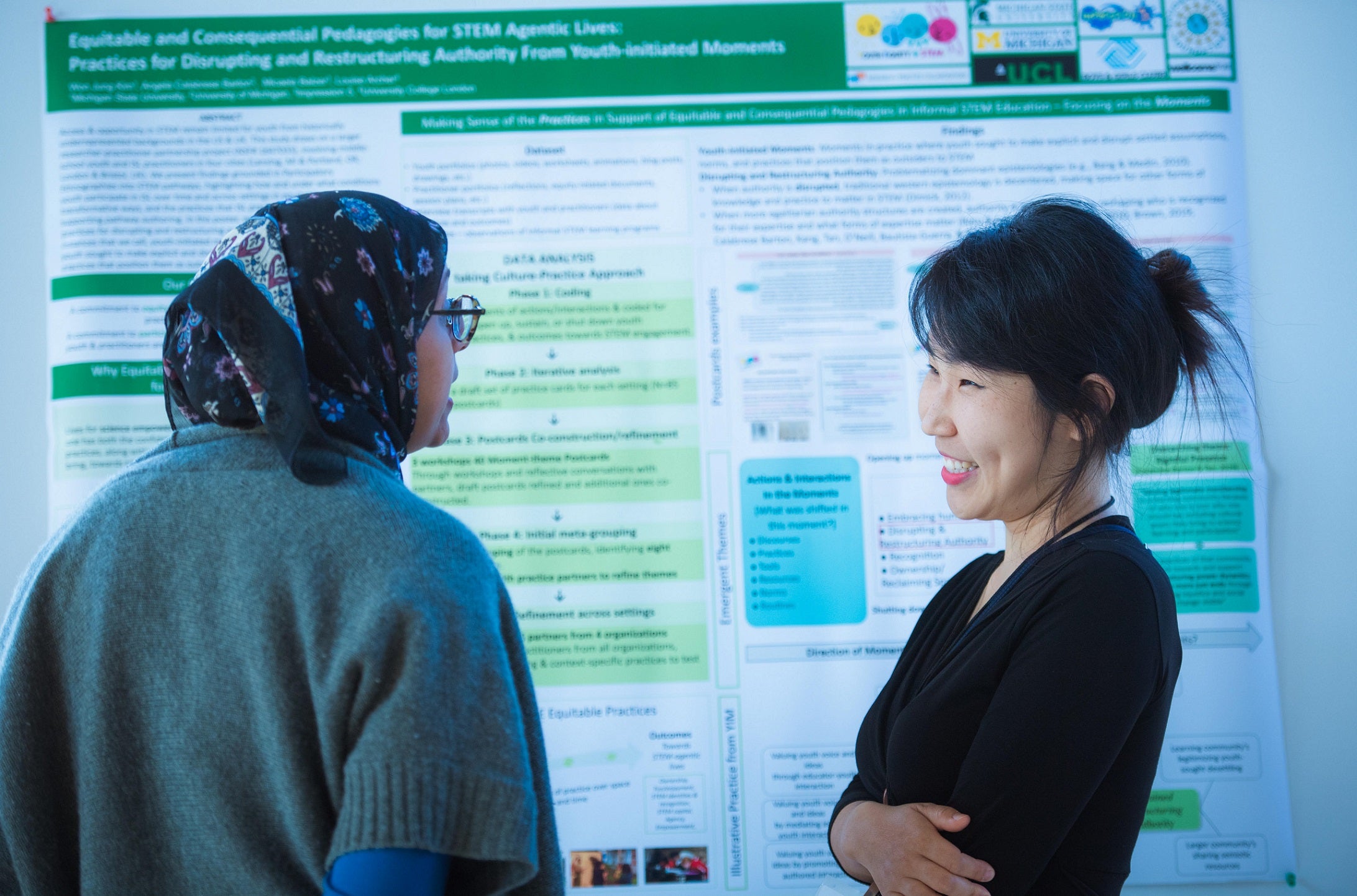 Two women talk in front of a scientific poster. One wears a headscarf, glasses, and a green sweater. The other woman with black hair smiles and listens.