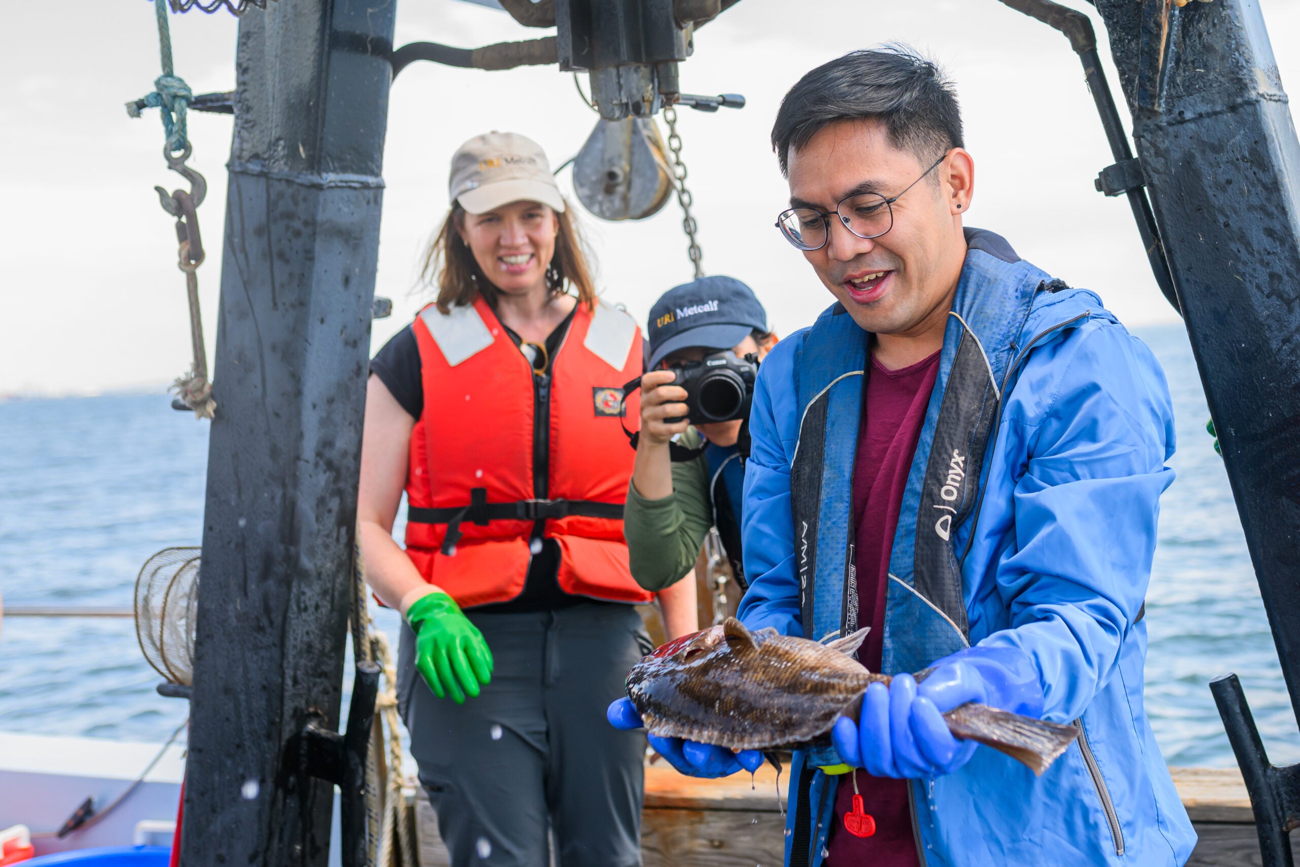 a scientist holds up a fish on a research boat while two people watch and take a photograph