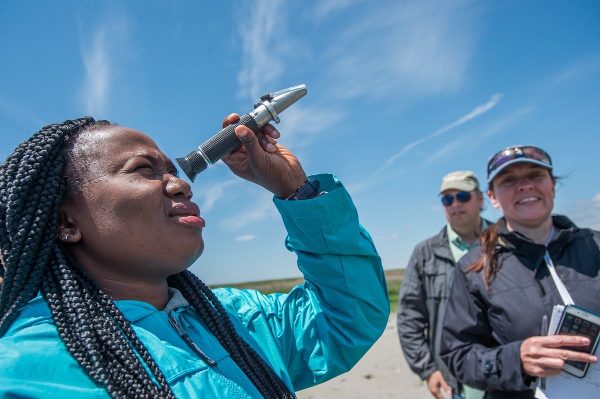 An African-American woman looks into a scientific device, there are two people in the background.