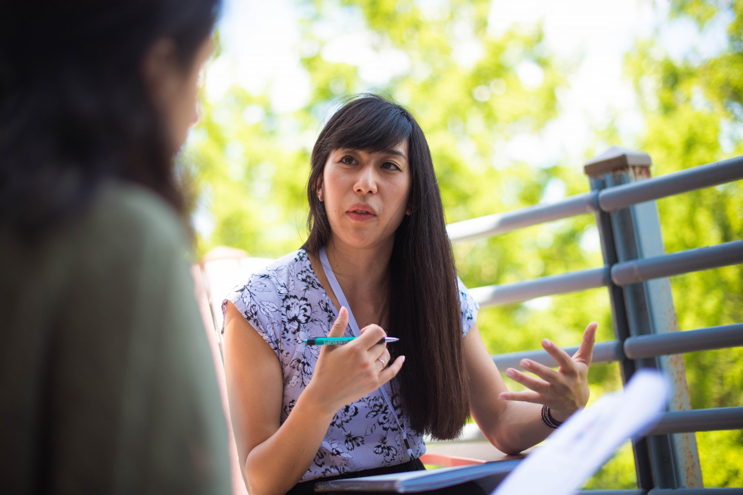 Photo of an Asian woman with long hair sitting outside. She is deep in discussion and gestures with her hands.