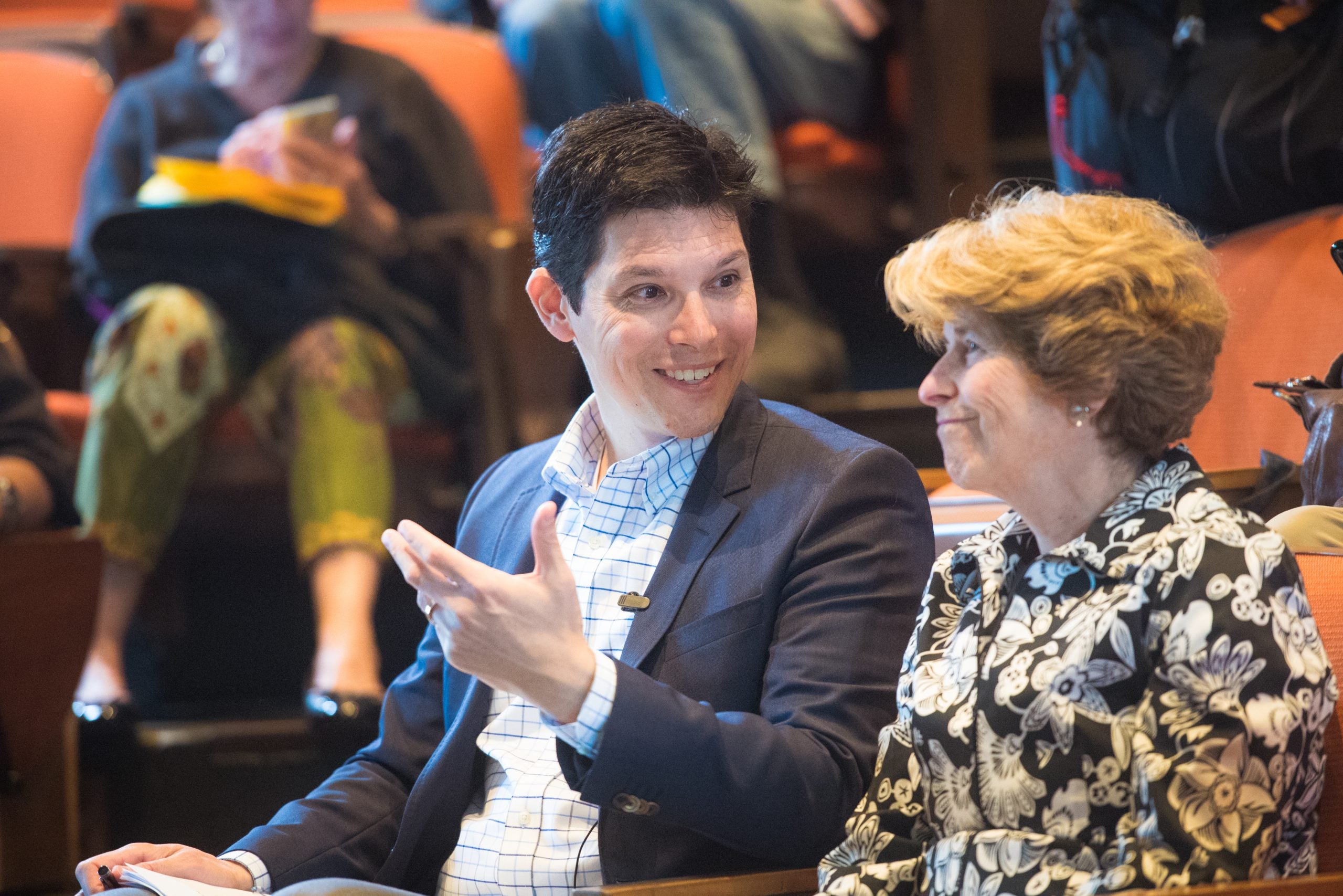 Two people are seated in an auditorium, talking. A light skinned man with short black hair gestures while smiling as a light-skinned woman with blond hair listens.