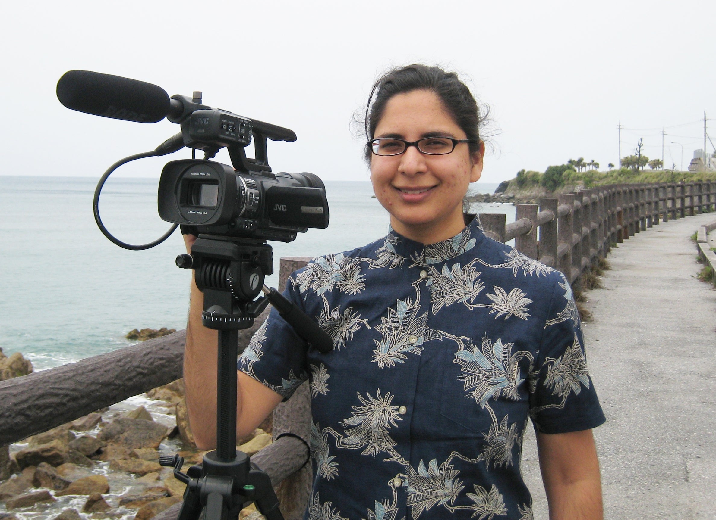 Photo of woman with black hair and glasses standing behind a video camera. There is a fence and coastal water in the background.