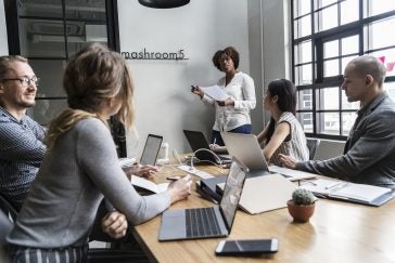 Business meeting setting with a Black woman standing next to a whiteboard and talking to two woman and two men sitting at a table